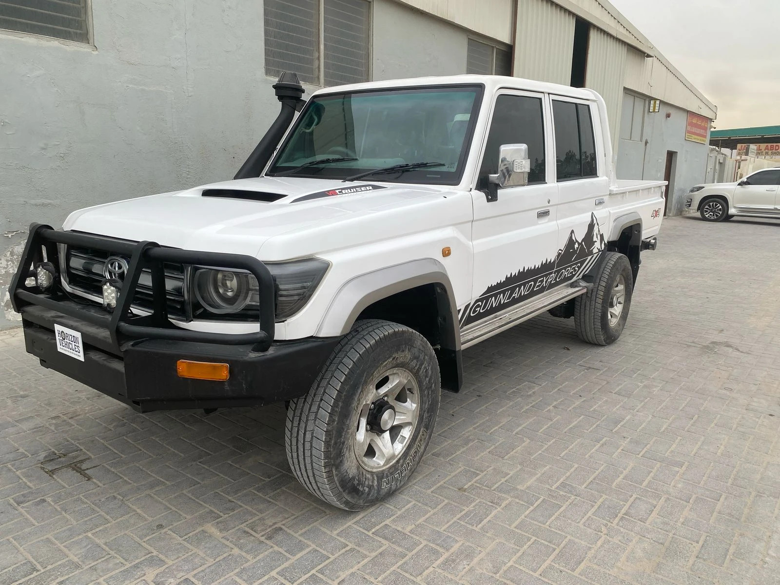 alt text: A white 2016 Toyota Land Cruiser double-cab pickup truck parked on a paved surface with buildings in the background.