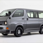 Front view of a beige Toyota HiAce Vanace Type 1 with dual round headlights, retro bumper, and chrome wheels.