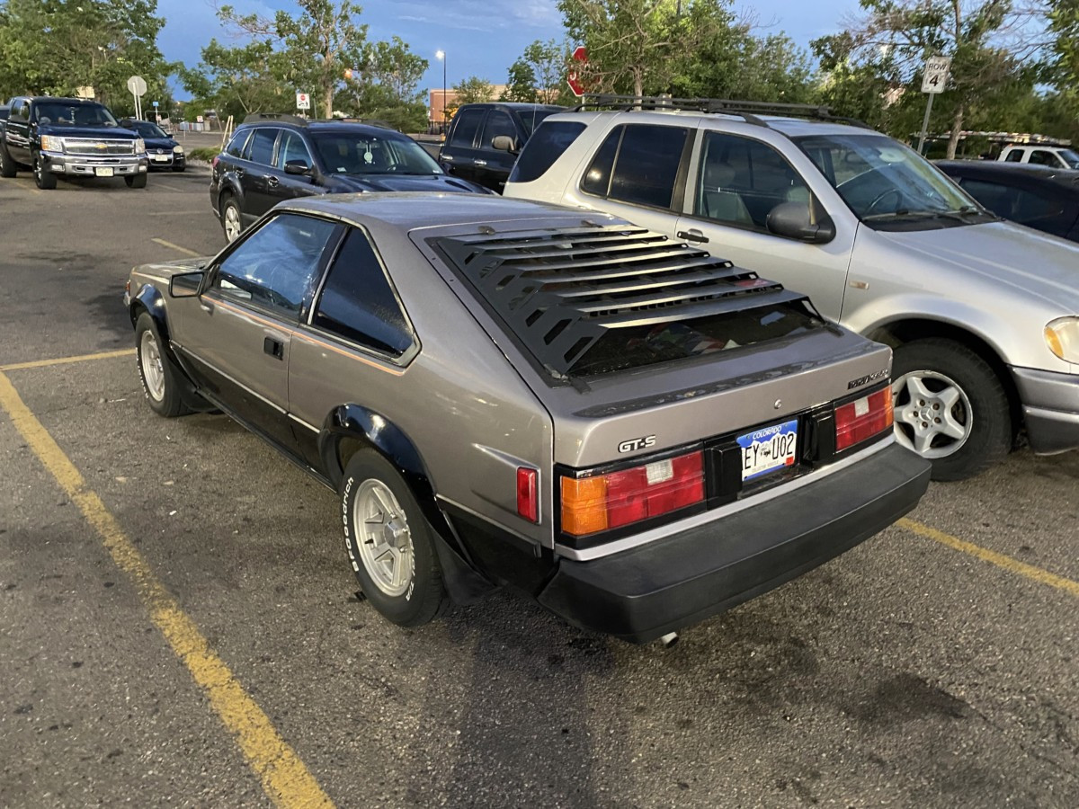 Front quarter view of a red 1985 Toyota Celica GT-S parked on a city street, showcasing its pop-up headlights and sporty design.