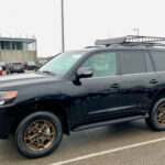 Midnight black 2021 Toyota Land Cruiser Heritage Edition parked in front of a Toyota dealership, showcasing its side profile and OEM running boards.