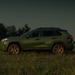 Side profile of army green 2024 Toyota RAV4 Hybrid parked on grass with trees in the background at nightfall.