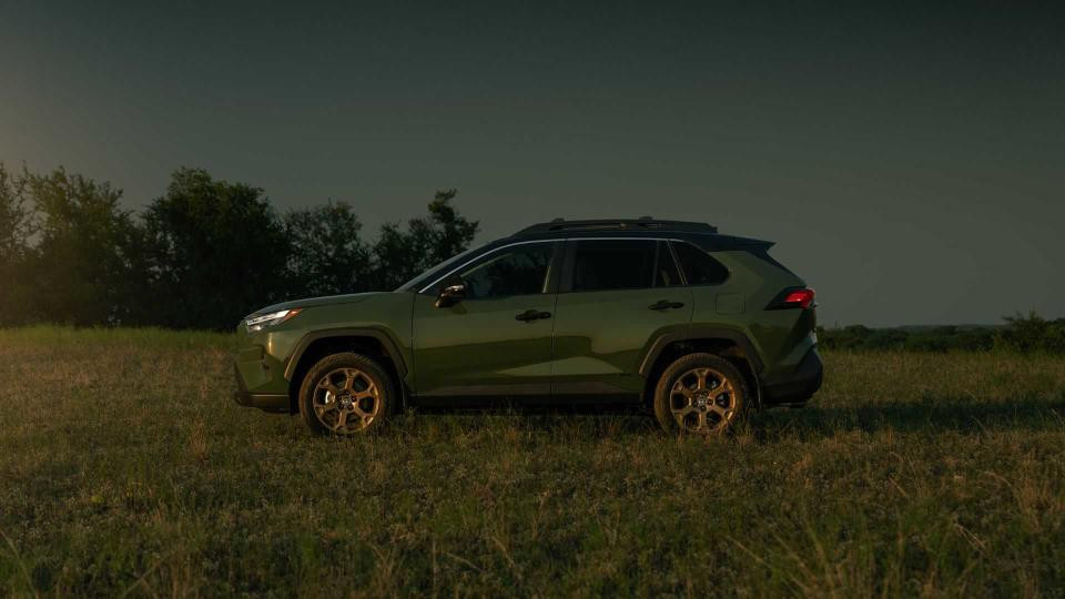 Side profile of army green 2024 Toyota RAV4 Hybrid parked on grass with trees in the background at nightfall.